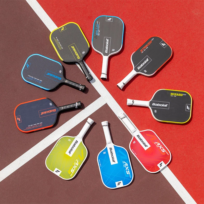 A collection of Babolat pickleball paddles in various colors and models arranged in a circular pattern on a red pickleball court background.